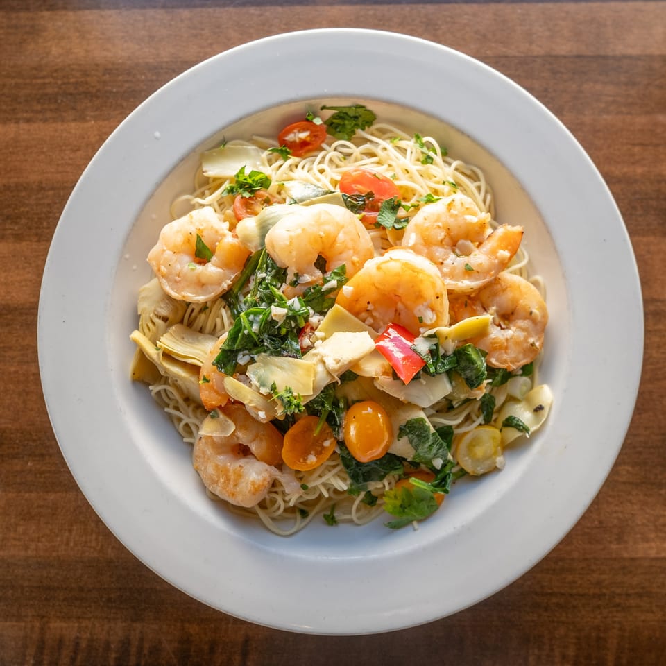 Plate of shrimp and angel hair pasta topped with spinach, artichokes, tomatoes, peppers, and herbs on a wooden table.