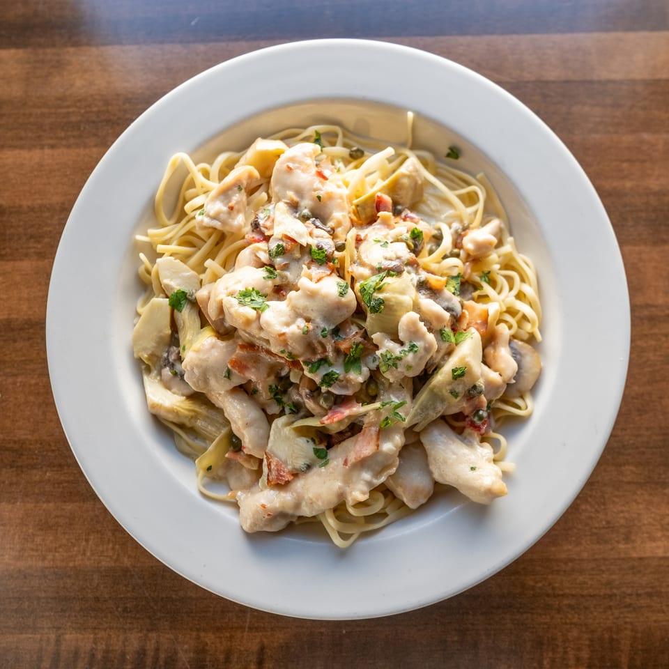Creamy chicken linguine with mushrooms, artichokes, tomatoes, capers, and parsley served on a white plate.
